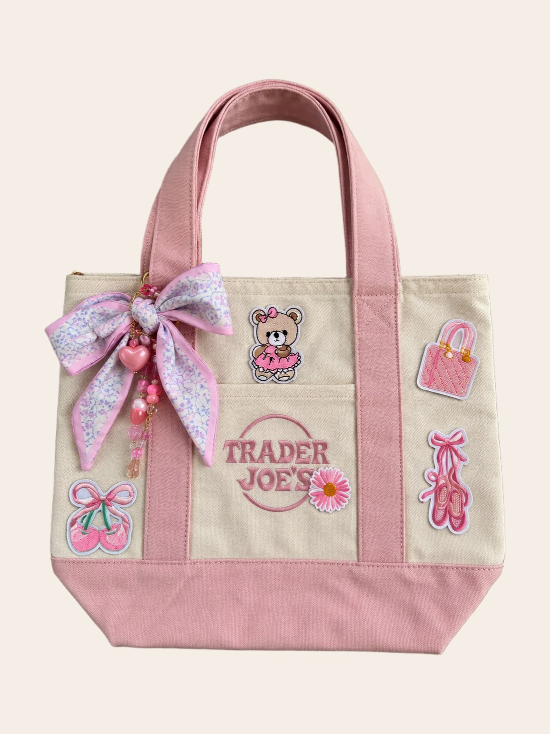 A cream and pink Trader Joe's mini tote with a teddy bear patch, a floral silk bow with heart charms, cherries, a pink purse patch and ballet slippers.